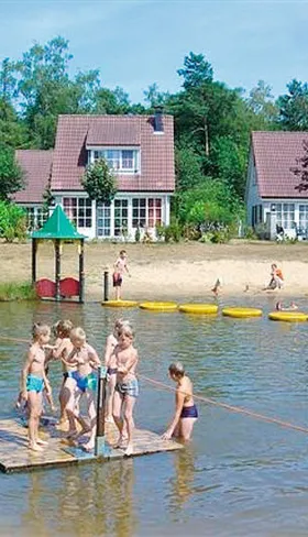 Kinderen spelen op een houten vlot in de zwemvijver van Vakantiepark Hellendoorn in Overijssel, met vakantiehuisjes op de achtergrond.