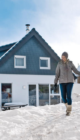 Een familie wandelt hand in hand door de sneeuw naar hun vrijstaande chalet op vakantiepark Landal Winterberg, met een kind op een slee.