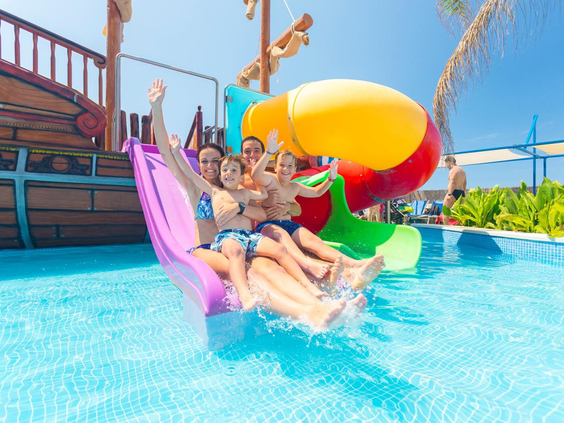 Een lachende familie met kinderen glijdt samen van de brede waterglijbaan in het zwembad van Royal Son Bou op Menorca.