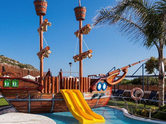 Groot houten piratenschip speeltoestel met een gele glijbaan in het kinderbad van familiehotel Royal Son Bou op Menorca.