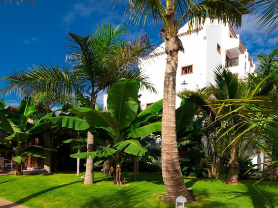 De weelderige tropische tuin van Parque Santiago III met palmbomen, bananenplanten en een groen gazon bij de witte appartementengebouwen.