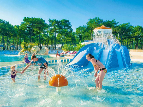 Peuter en ouders spelen met een spuitende fontein in het ondiepe, kindvriendelijke peuterbad van camping Atlantic Club Montalivet.
