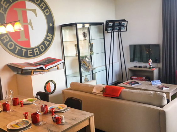 De woonkamer van de speciale Feyenoord Lodge op Bad Hoophuizen, ingericht met clublogo, een prijzenkast en andere fan-items.