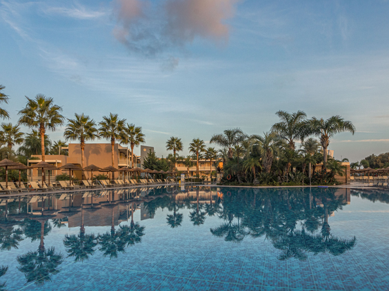 Het grote zwembad van Blue Lagoon Resort op Kos, met palmbomen die prachtig weerspiegelen in het kalme water tijdens zonsondergang.
