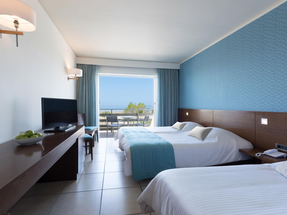 Een ruime familiekamer in het Blue Lagoon Resort op Kos, met drie opgemaakte bedden, een blauwe accentmuur en een deur naar het balkon.