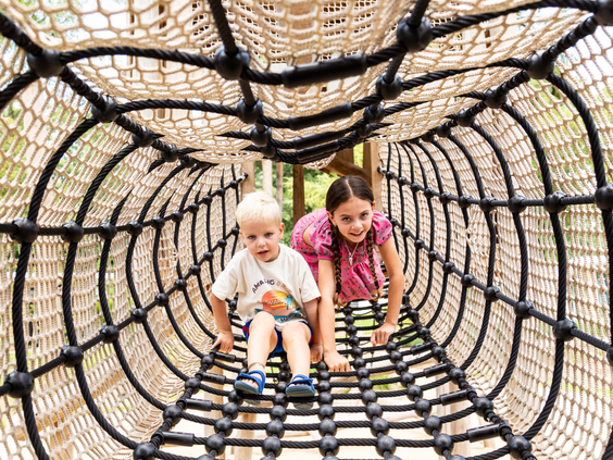 Twee kinderen klimmen door een tunnel van netten in de buitenspeeltuin van Bospark Ede op de Veluwe.