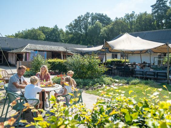 Een familie geniet van de lunch aan een tuintafel op het zonnige terras van Bospark Lunsbergen, met uitzicht op de speeltuin.