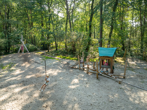 Overzicht van de natuurlijke bosspeeltuin van Bospark Lunsbergen, met een kabelbaan, een houten klimtoestel en veel speelzand, omgeven door hoge bomen.