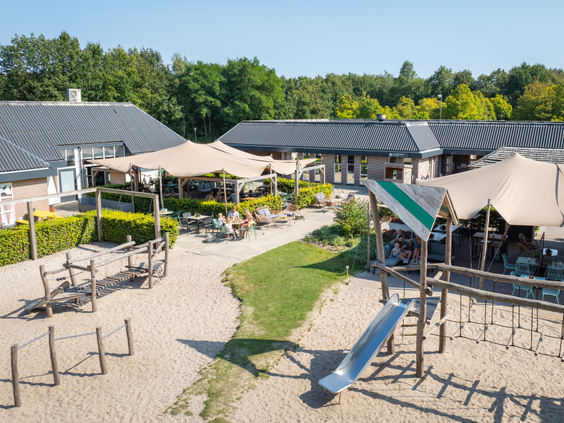 Zonnig terras met uitzicht op de speeltuin met zand en een glijbaan bij de centrumvoorzieningen van kindvriendelijk vakantiepark Bospark Lunsbergen.