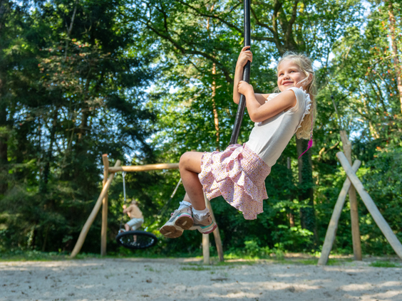 Een lachend meisje zoeft met de kabelbaan door de bosspeeltuin van Bospark Lunsbergen, omringd door bomen en andere houten speeltoestellen.