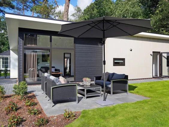 Het ruime terras van een modern vakantiehuis op Bospark Markelo, ingericht met een luxe loungeset en een grote parasol op een groen gazon.