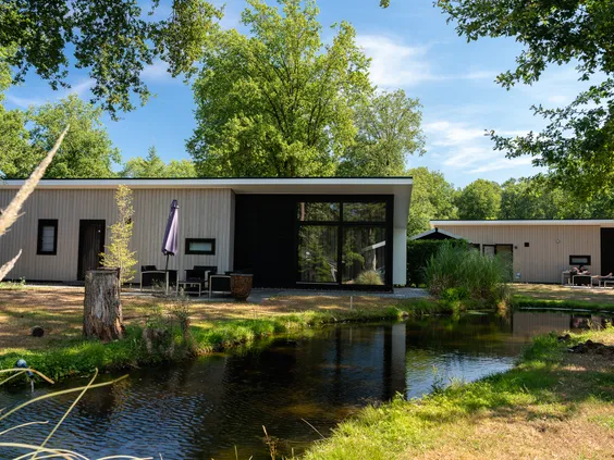Een modern, lichtgekleurd chalet met grote ramen en een terras direct aan de waterkant op het bosrijke Bospark Markelo.
