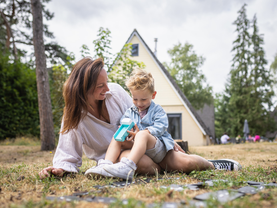 Een moeder en haar zoontje zitten op een kleed in het gras voor hun bungalow te spelen met een bellenblaas op vakantiepark De Katjeskelder.