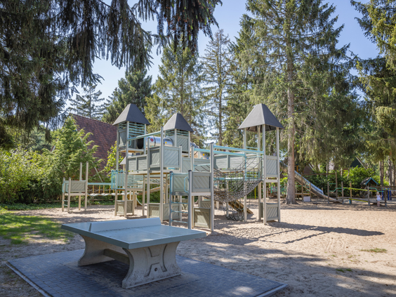 Een groot houten speelkasteel met glijbanen en een tafeltennistafel in de avontuurlijke buitenspeeltuin van De Katjeskelder, omgeven door bomen.