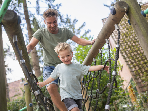 Een vader helpt zijn zoontje balanceren op een wiebelende touwbrug in de avontuurlijke speeltuin van Bungalowpark De Katjeskelder.