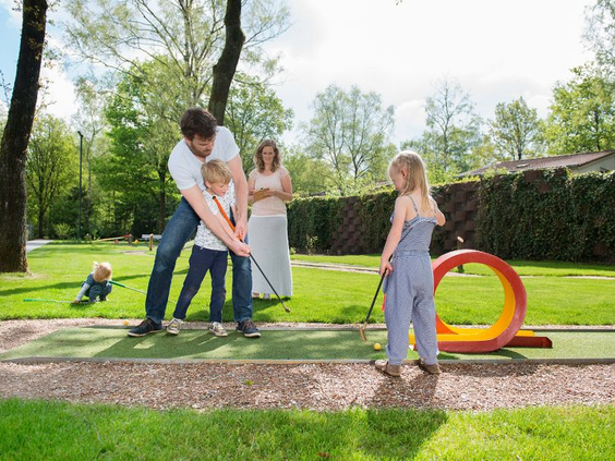 Een gezin met jonge kinderen speelt midgetgolf op de midgetgolfbaan van Bungalowpark Hoenderloo, gelegen in het groen.