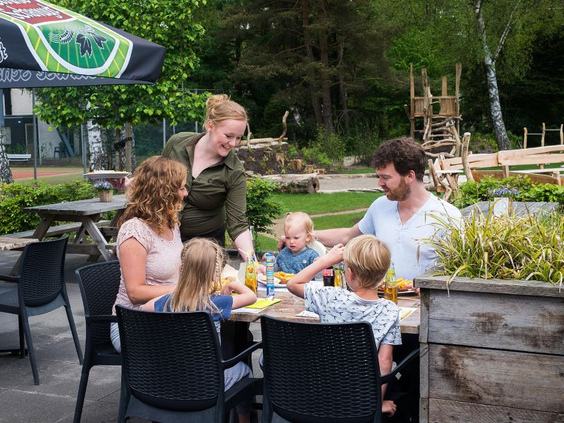Een gezin met kinderen geniet van een drankje op het terras, direct naast de buitenspeeltuin van Bungalowpark Hoenderloo.