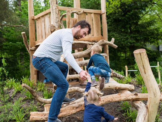 Een vader helpt zijn kinderen met klimmen op een natuurlijk, houten speeltoestel in het speelbos van Bungalowpark Hoenderloo.