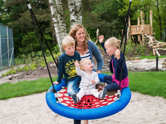 Een moeder lacht terwijl ze met haar drie kinderen op een grote, blauwe nestschommel in de speeltuin van Bungalowpark Hoenderloo zit.