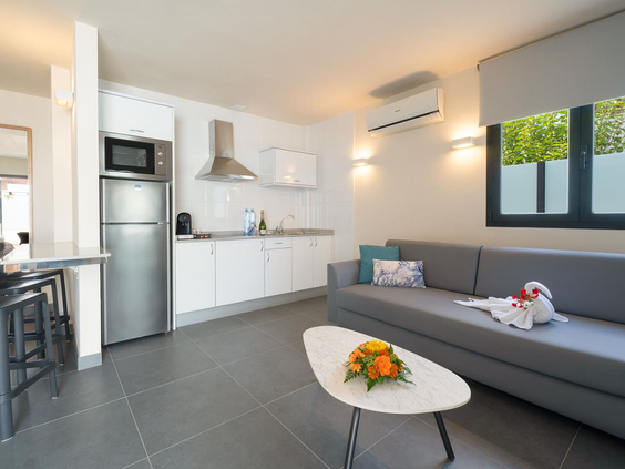 Moderne woonkamer met open keuken in een bungalow van Relaxia Lanzasur Club, voorzien van een grijze hoekbank, eethoek en koelkast.