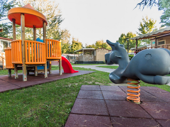 Een veilig, omheind speeltuintje met een houten speeltoestel en een wipkip op een zachte ondergrond, ideaal voor peuters en kleuters op Camping Altomincio.