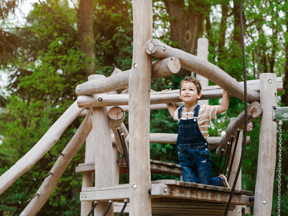 Een jongen in een tuinbroek klimt behendig over een houten speeltoestel in het bos op Camping Beekse Bergen.