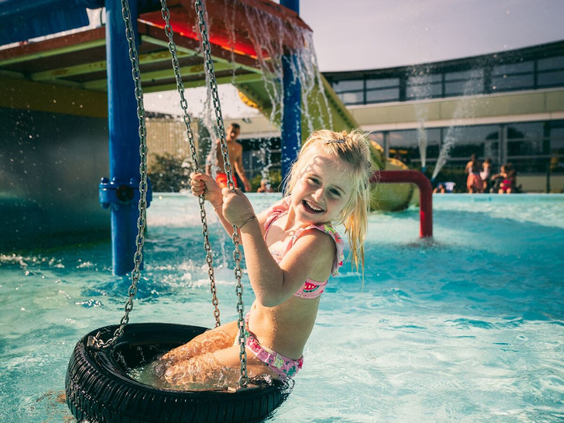 Meisje schommelt in een band onder een waterval in de waterspeeltuin van het zwembad op Camping de Schatberg.