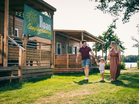 Gezin wandelt hand in hand voor hun houten stacaravan op een grasveld van Camping de Schatberg.