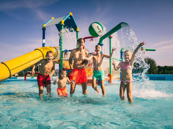 Groep vrolijke kinderen wordt nat onder de grote omkiepende emmer in de waterspeeltuin van Camping de Schatberg.