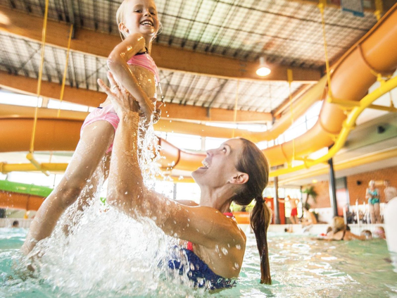 Moeder tilt haar lachende dochter op uit het water in het binnenzwembad van Camping de Schatberg.