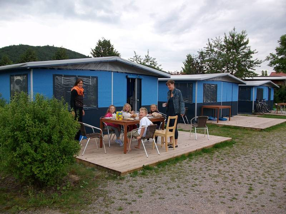 Een familie zit gezellig aan een tafel op het terras voor hun blauwe houten trekkershut op Knaus Campingpark Eschwege.