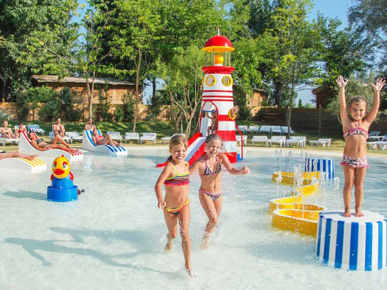 Kinderen rennen en spelen vrolijk in de ondiepe waterspeeltuin met een vuurtoren en andere speeltoestellen op Camping Le Sérignan Plage.