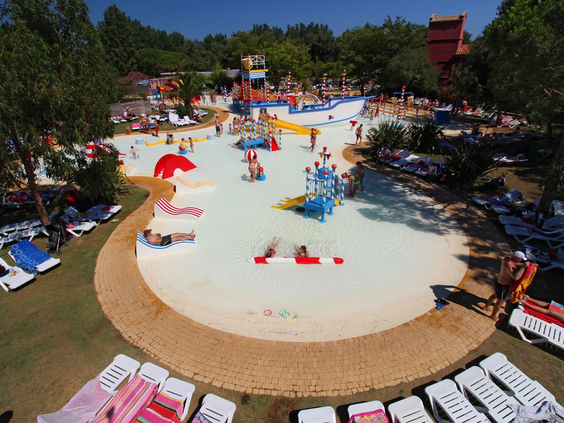 Overzicht van boven van de grote, ondiepe waterspeeltuin met speeltoestellen, omringd door ligbedjes op Camping Le Sérignan Plage.