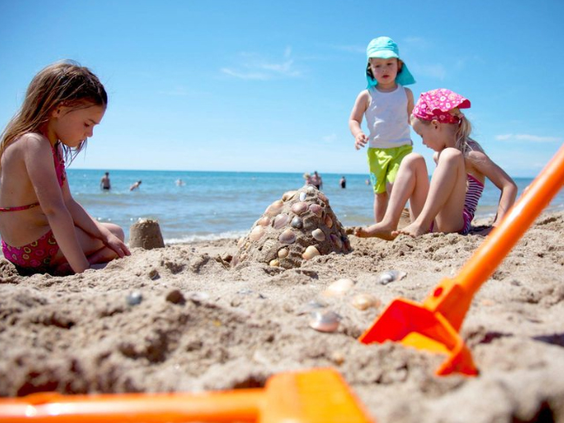 Drie jonge kinderen bouwen een groot zandkasteel met schelpen op het brede zandstrand bij Camping Le Sérignan Plage.