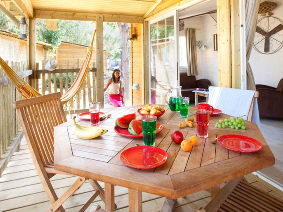 Gedeeltelijk gedekte houten eettafel met vers fruit en een hangmat op de veranda van een stacaravan op Camping Le Sérignan Plage.