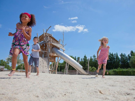 Drie kinderen rennen en springen vrolijk in de grote zandspeeltuin met een houten klimtoestel en glijbaan op Camping Le Sérignan Plage.