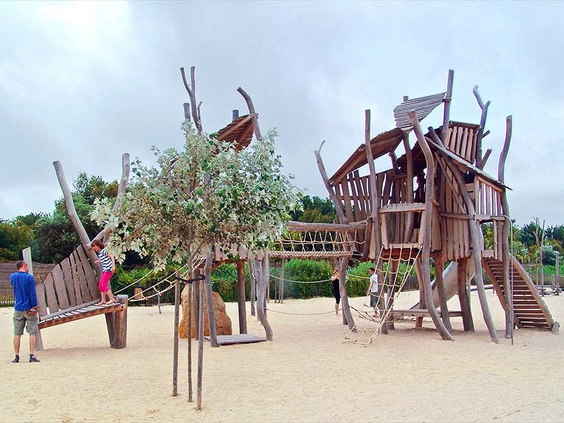 Groot avontuurlijk houten speeltoestel in de vorm van een schip, gelegen in een zanderige speeltuin op Camping Le Sérignan Plage.