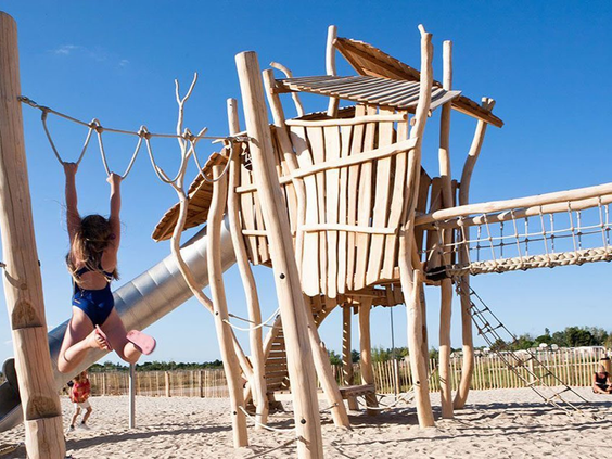 Meisje klimt behendig over een touwbrug van het houten klimrek in de avontuurlijke speeltuin van Camping Le Sérignan Plage.