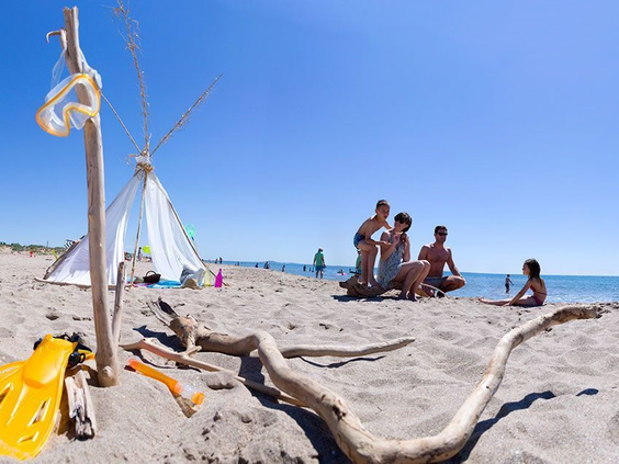 Een gezin zit gezellig in het zand aan de rand van de zee op het strand van Camping Le Sérignan Plage, met een zelfgemaakte tent van drijfhout.