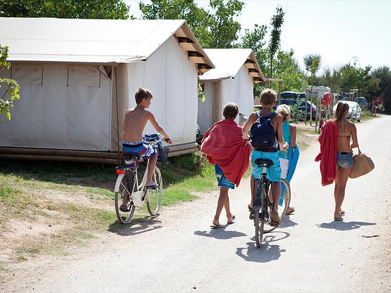 Een groep kinderen en tieners met handdoeken en fietsen loopt over een pad langs safaritenten op Camping Le Sérignan Plage, op weg naar het strand.