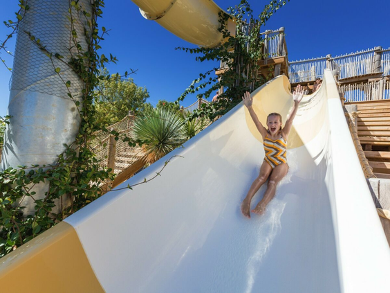 Kind met de armen in de lucht glijdt met plezier van een wit-gele waterglijbaan op Camping Le Sérignan Plage.