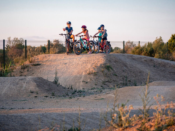 Drie kinderen met fietsen en helmen staan boven op een heuvel van de zanderige pumptrack of crossbaan op Camping Le Sérignan Plage.
