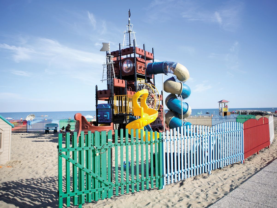 Groot speeltoestel in de vorm van een kinderspeelkasteel, gelegen in het zand van het strand op Camping Union Lido, omheind met een kleurrijk hek.