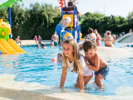 Twee blije kinderen spelen samen in het ondiepe water van het kinderbad op Camping Union Lido, met kleurrijke speeltoestellen op de achtergrond.