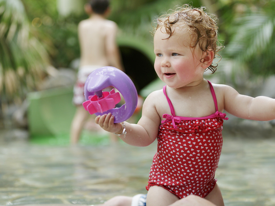 Een lachende dreumes in een rood badpakje speelt met een bal in het ondiepe water van het subtropische zwemparadijs Aqua Mundo bij Center Parcs Bispinger Heide.