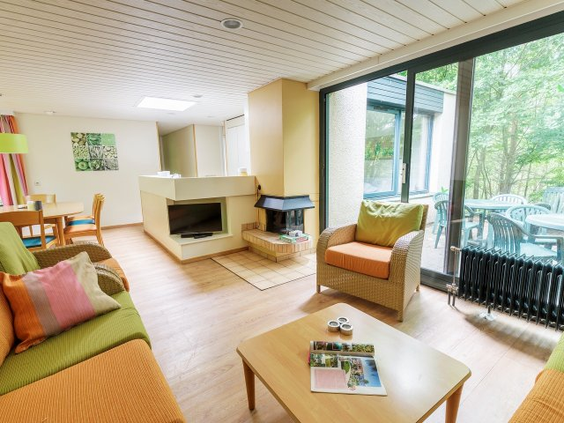 Gezellige woonkamer van een cottage in Center Parcs Bispinger Heide, met een comfortabele zithoek, open haard en grote ramen met uitzicht op het terras en het bos.