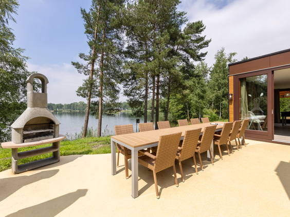 Een groot terras met een lange eettafel en een barbecue van een luxe cottage aan het meer bij Center Parcs De Kempervennen.