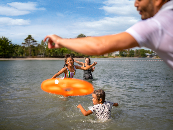 Een gezin heeft waterpret in het meer bij Center Parcs De Kempervennen, spelend met een oranje zwemband op een zonnige dag.