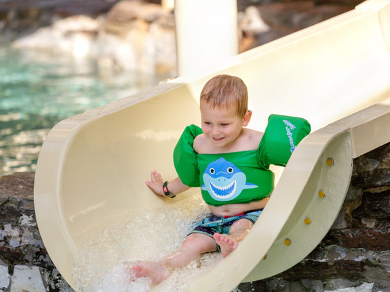 Een peuter met zwemvleugels glijdt lachend van een kleine waterglijbaan in het Kinder-doe-bad van de Aqua Mundo op De Kempervennen.