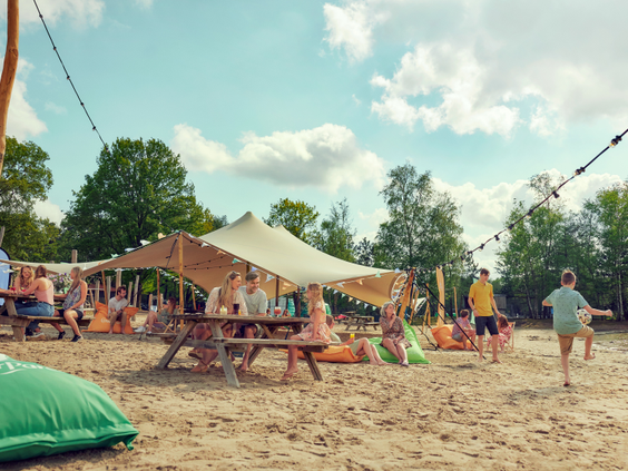 Mensen ontspannen op het zandstrand bij een gezellig strandpaviljoen met een grote tent en zitzakken aan het meer van Center Parcs De Kempervennen.